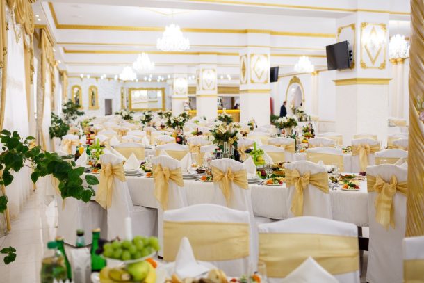 An elegant banquet hall set up for a wedding reception, featuring long tables dressed in white linens and chairs adorned with gold satin bows. The room is brightly lit by grand crystal chandeliers, showcasing intricate gold wall moldings and beautiful floral centerpieces running down the lengths of the tables.