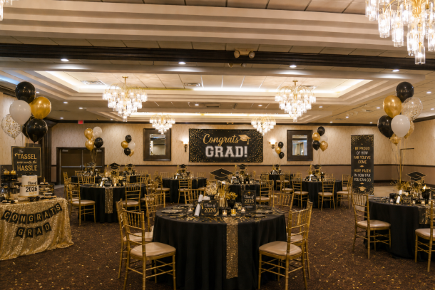 A beautifully decorated banquet room at Club Venetian set up for a 2026 graduation party. The round tables are dressed in black linens with gold chairs, and a large "Congrats GRAD!" banner hangs on the back wall flanked by black, gold, and white balloon arches.