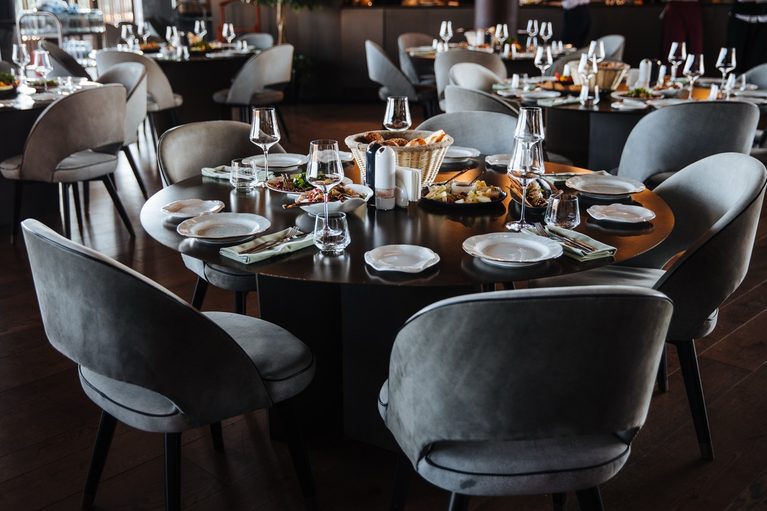 An elegantly arranged round banquet dining table set for guests with white china, polished cutlery, wine glasses, folded napkins, and light gray velvet armchairs.
