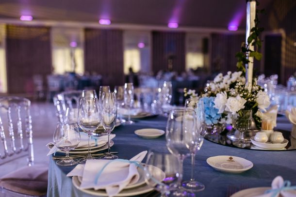An elegant banquet dining table setup with blue glassware, folded white napkins, and polished dinnerware in a brightly lit event space.